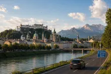 Golden-hour view of Salzburg with a river running through the city, historic domed churches and a hilltop fortress overlooking the skyline, mountains in the background, and a roadside sign indicating a route toward Brussels (850 km) beside a car on a scenic highway.