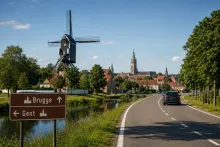 A scenic countryside road in Flanders, Belgium, showing a car driving along a quiet route between Ghent and Bruges. In the foreground, directional road signs point toward Brugge and Gent beside a canal. The landscape features a traditional windmill, historic village houses, green trees, and a distant church tower under a clear blue sky, illustrating a road trip route through rural Belgium.