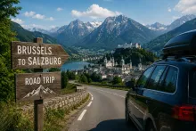 A scenic alpine road trip scene featuring a black SUV driving along a winding mountain road toward Salzburg. In the foreground, a rustic wooden sign reads “Brussels to Salzburg Road Trip.” The background shows a picturesque European town by a lake, surrounded by lush green hills and dramatic snow-capped mountains under a bright blue sky.