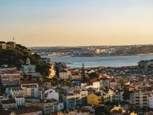 Panoramic view of Lisbon skyline with colorful hillside buildings, historic rooftops, the Tagus River, and the 25 de Abril Bridge under warm sunset light.