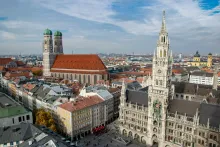 Panoramic view of Munich city center with historic architecture, church towers, and the Bavarian Alps visible in the distance.