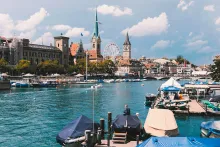 Panoramic view of Zurich, Switzerland featuring the Limmat River, historic old town buildings, church towers, waterfront promenades, and the surrounding alpine landscape under clear skies.