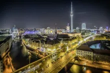 Panoramic view of Berlin, Germany, featuring the historic city center, the Fernsehturm rising above the skyline, elegant government buildings, church domes, modern architecture, and the River Spree winding through the city under a clear sky.