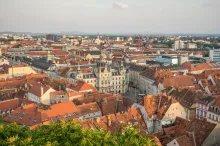 Panoramic view of Graz, Austria, featuring the red rooftops of the UNESCO-listed Old Town, Schlossberg Hill with the iconic Clock Tower, the Mur River, and surrounding Alpine foothills beneath a clear blue sky.