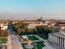 Panoramic view of Munich, Germany, featuring the historic Marienplatz with the Neues Rathaus, Gothic architecture, city rooftops, church towers, green parks, and the urban skyline of Bavaria’s capital under a clear blue sky.