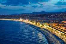 Panoramic view of Nice, France, featuring the turquoise Mediterranean Sea, the Promenade des Anglais, colorful Old Town buildings, Castle Hill, palm trees, and the dramatic coastal landscape of the French Riviera under a clear blue sky.