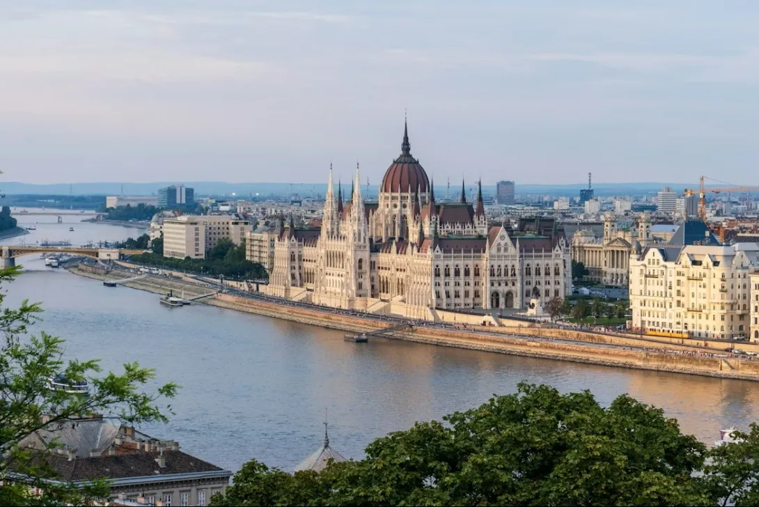 Panoramic view of the Hungarian Parliament Building along the banks of the Danube River in Budapest at sunset, with city skyline and riverside promenade visible.