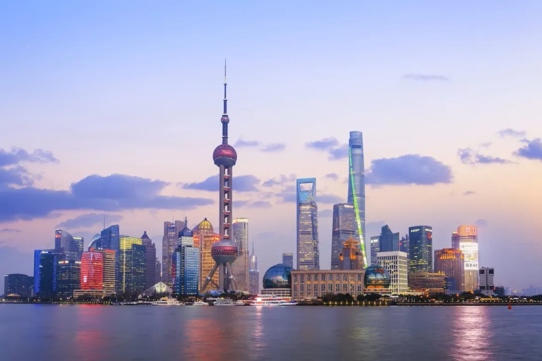 A panoramic view of Shanghai's futuristic Pudong skyline at dusk, featuring the Oriental Pearl TV Tower and Shanghai Tower, with their lights reflecting on the Huangpu River.