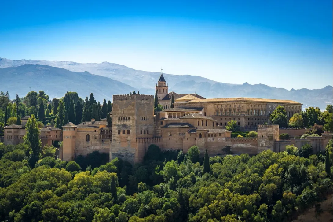 The Alhambra palace complex in Granada, Spain, featuring the ancient Moorish fortifications and the Palace of Charles V, set against the backdrop of the Sierra Nevada mountains under a clear blue sky.