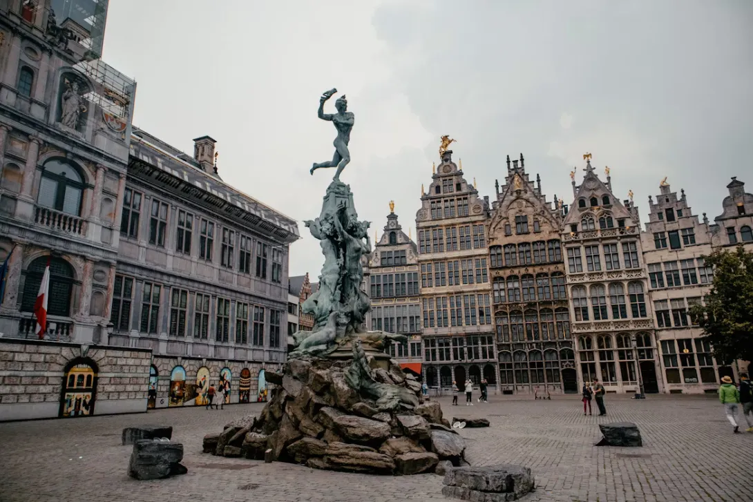 Panoramic view of Antwerp, Belgium featuring the historic old town, cathedral spire, riverside architecture along the Scheldt River, and colorful European rooftops under golden evening light.
