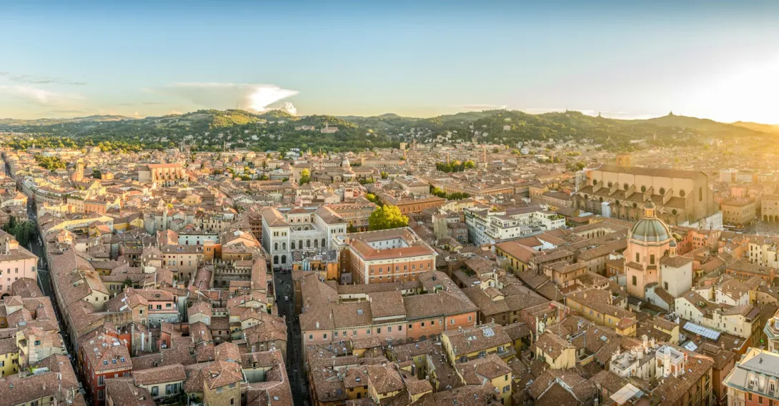 Panoramic view of Bologna city skyline with its terracotta rooftops, historic towers, and surrounding Emilia-Romagna hills under warm evening light.