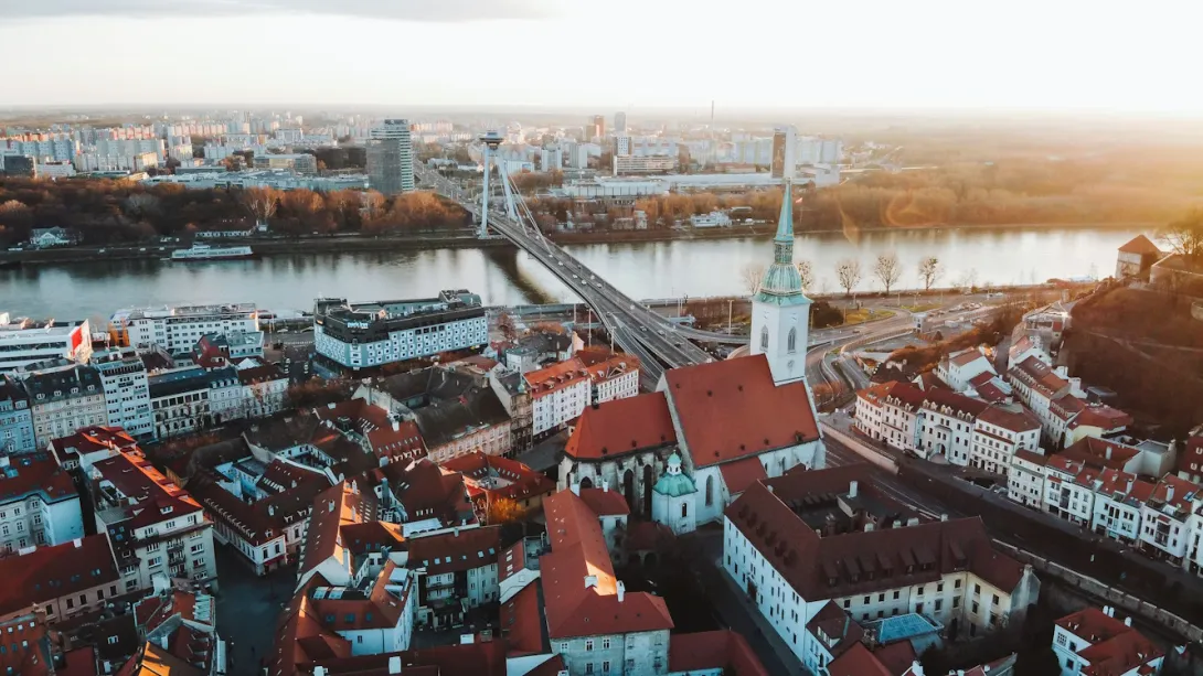 Panoramic view of Bratislava, Slovakia, featuring Bratislava Castle above the Danube River, historic old town rooftops, bridges, and the modern city skyline at sunset.