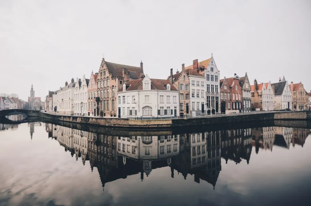 Panoramic view of Bruges with medieval canals, cobblestone streets, historic Flemish buildings, and church towers reflected in calm water under golden sunset light in Belgium.