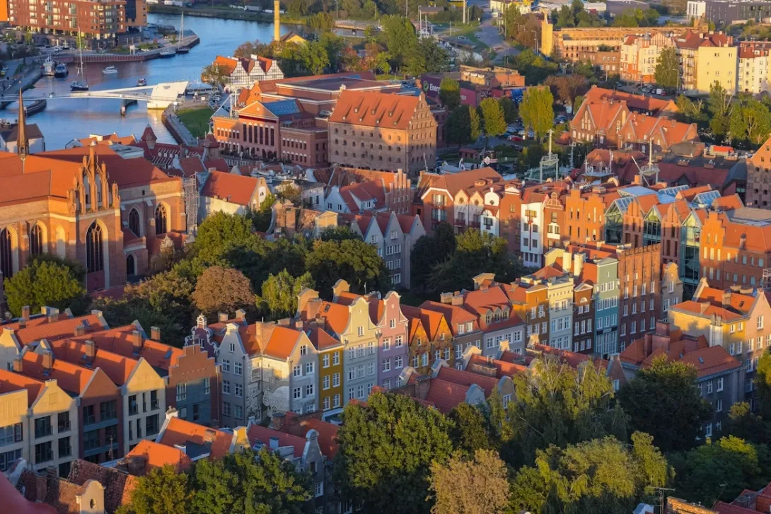 Panoramic view of Gdansk, Poland featuring colorful historic Hanseatic buildings along the Motława River, medieval cranes, and waterfront reflections under a clear sky at sunset.
