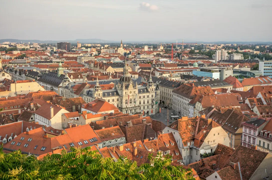 Panoramic view of Graz, Austria featuring red-tiled rooftops, historic old town architecture, the Mur River, and Schlossberg hill overlooking the city under clear alpine skies.