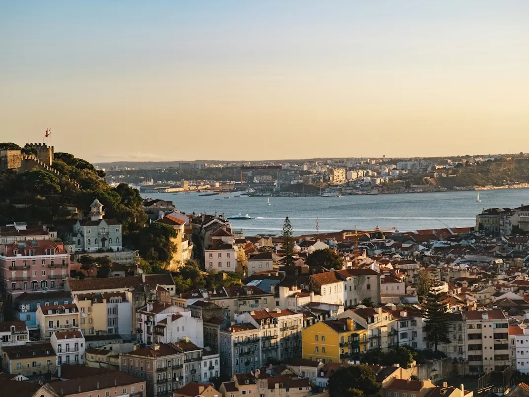 Panoramic view of Lisbon skyline with colorful hillside buildings, historic rooftops, the Tagus River, and the 25 de Abril Bridge under warm sunset light.