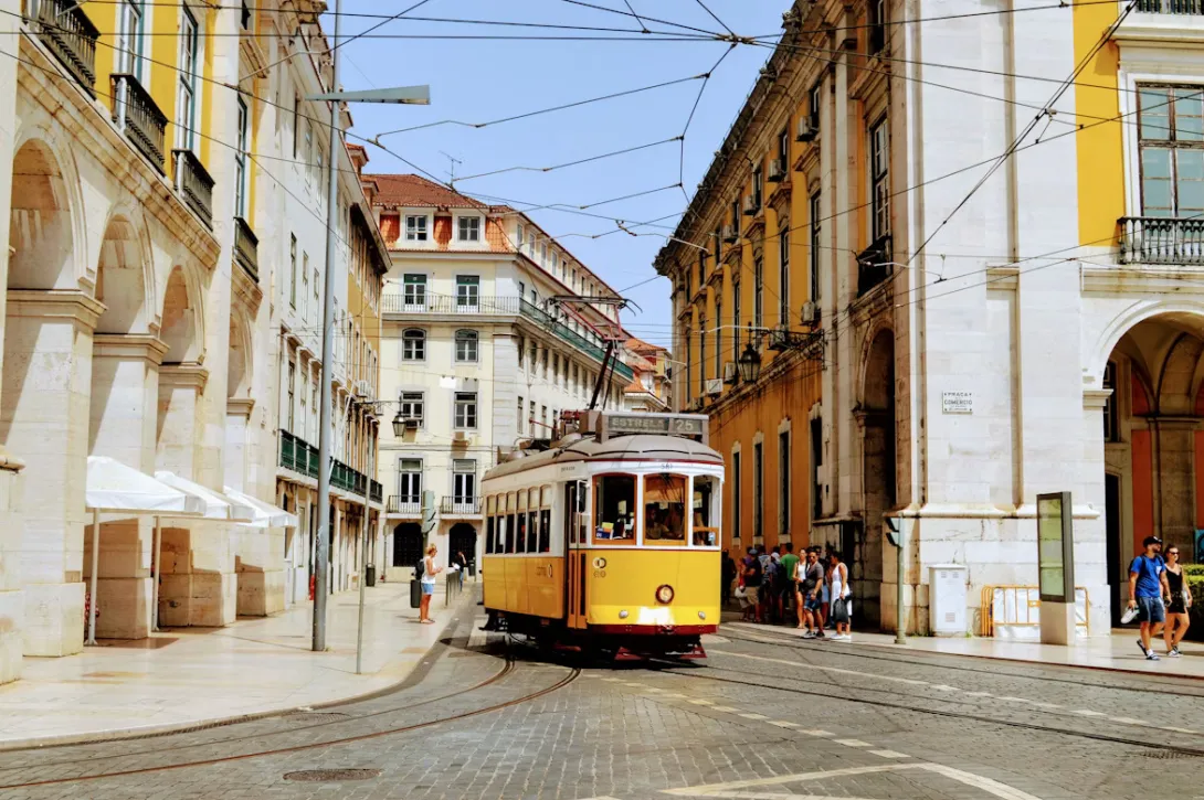 Panoramic view of Lisbon, Portugal at sunset with colorful historic buildings, red rooftops, tram lines, and the Tagus River stretching across the city skyline.