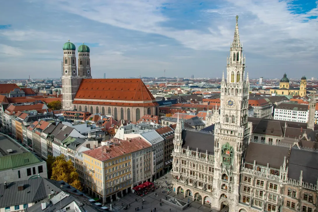 Panoramic view of Munich city center with historic architecture, church towers, and the Bavarian Alps visible in the distance.
