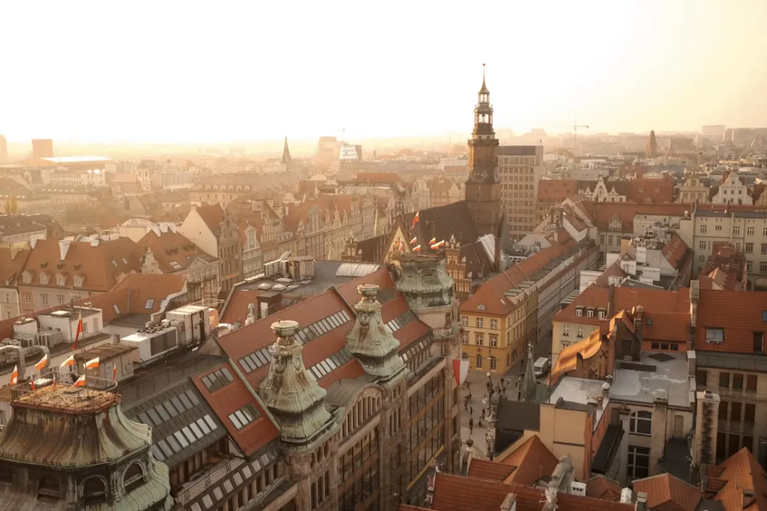Panoramic view of Wroclaw, Poland featuring colorful market square buildings, historic church towers, cobblestone streets, and the Oder River winding through the city at sunset.