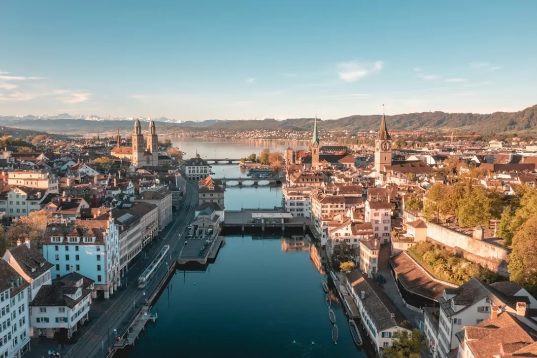 Panoramic view of Zurich city skyline with Lake Zurich, historic church towers, and the Swiss Alps visible in the distance.