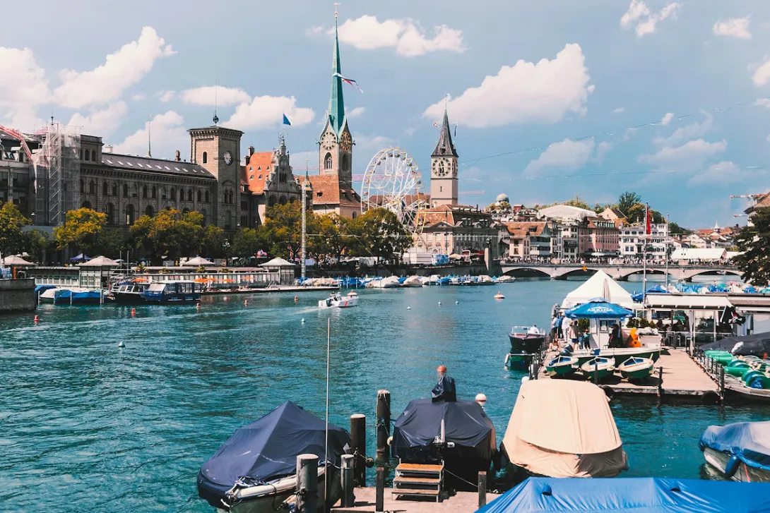 Panoramic view of Zurich, Switzerland featuring the Limmat River, historic old town buildings, church towers, waterfront promenades, and the surrounding alpine landscape under clear skies.