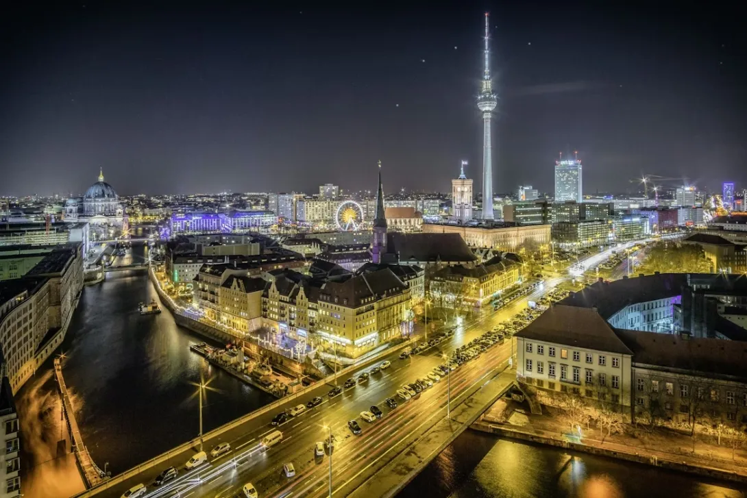 Panoramic view of Berlin, Germany, featuring the historic city center, the Fernsehturm rising above the skyline, elegant government buildings, church domes, modern architecture, and the River Spree winding through the city under a clear sky.