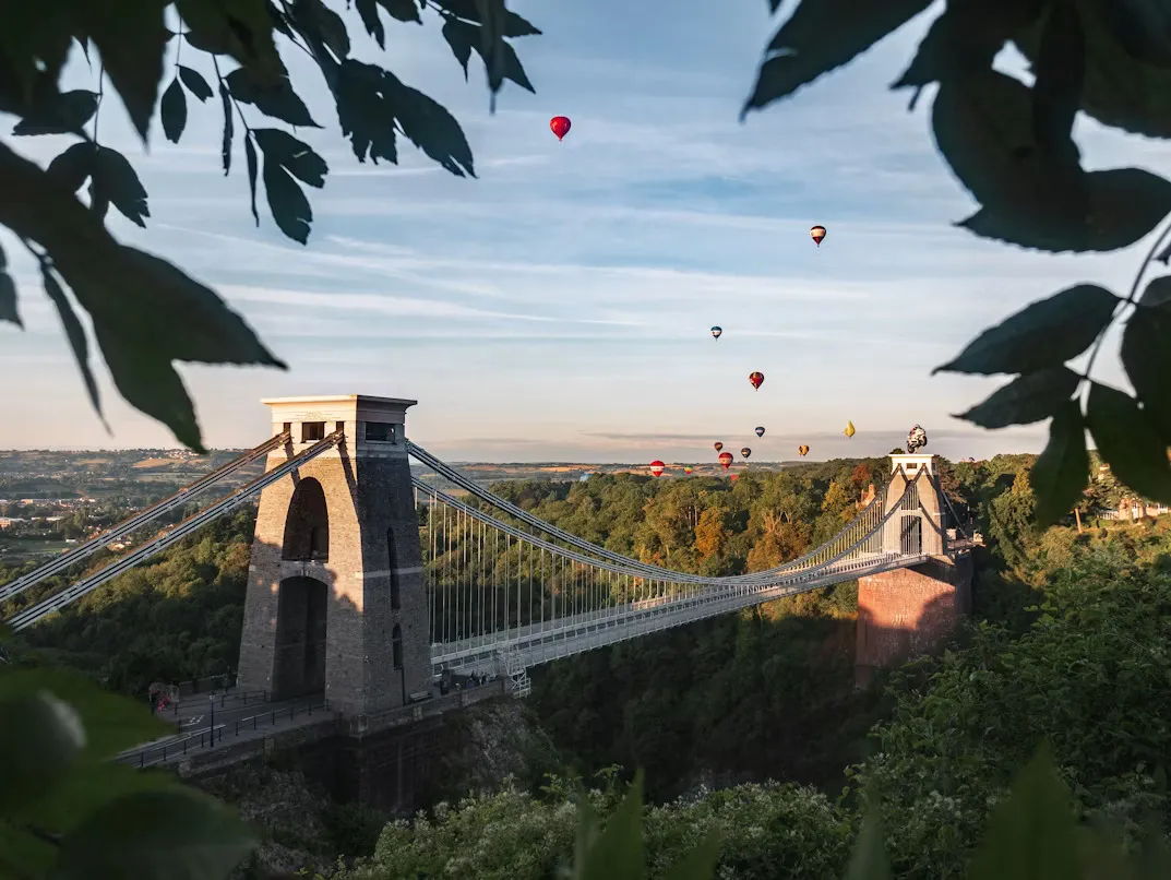 Panoramic view of Bristol, England, featuring the Clifton Suspension Bridge, the River Avon gorge, historic Georgian buildings, and green rolling hills under soft cloudy skies.