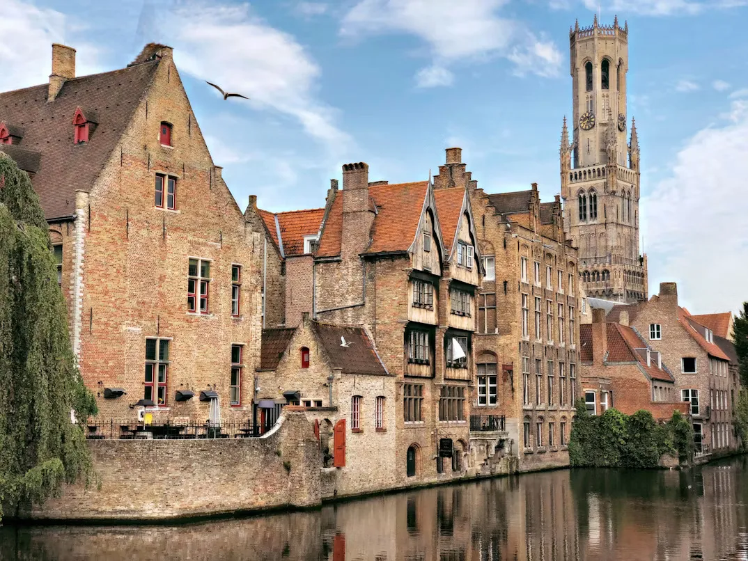 Panoramic view of Bruges, Belgium, featuring medieval brick buildings, historic church towers, winding canals, stone bridges, and traditional Flemish architecture reflected in the water under a clear sky.
