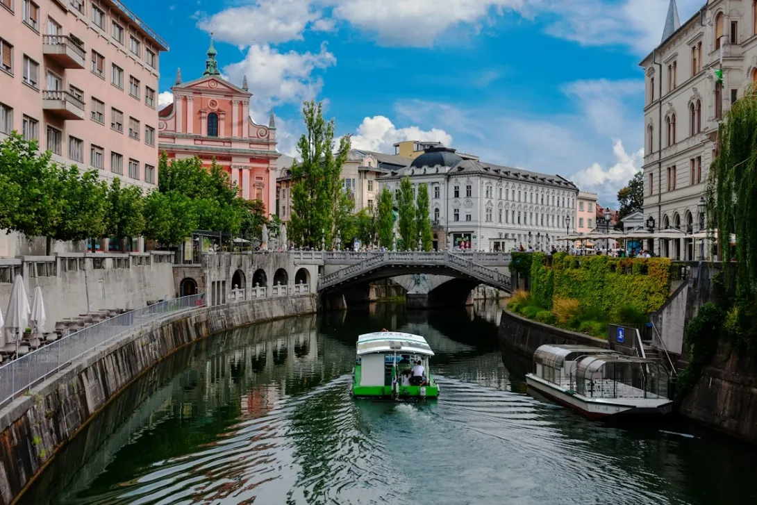 Panoramic view of Ljubljana, Slovenia, featuring the Ljubljanica River, colorful historic buildings, red-tiled rooftops, elegant bridges, Ljubljana Castle rising above the city, and surrounding green hills under a clear sky.