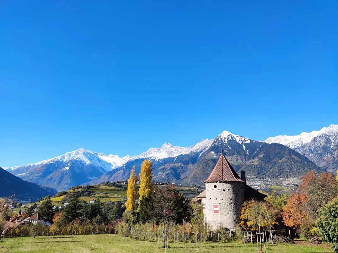 Panoramic view of Merano, Italy, featuring elegant Alpine architecture, the Passer River, lush gardens, and surrounding mountain peaks beneath a clear South Tyrolean sky.