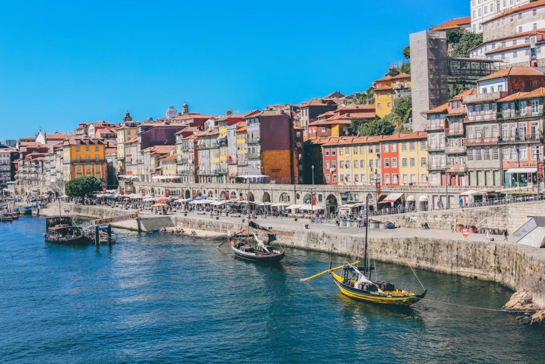 Panoramic view of Porto, Portugal, featuring colorful riverside buildings along the Douro River, historic church towers, terracotta rooftops, traditional architecture, and the iconic Dom Luís I Bridge under a bright blue sky.