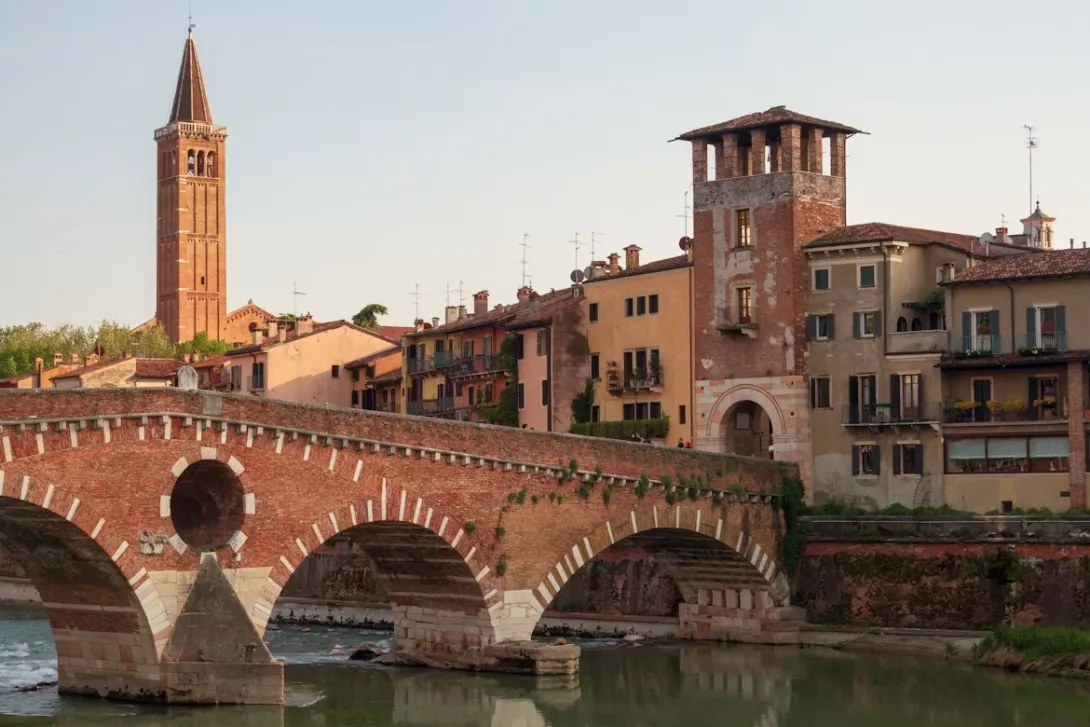 Panoramic view of Verona, Italy, featuring historic terracotta rooftops, medieval towers, the Adige River winding through the city, elegant bridges, and centuries-old Italian architecture beneath a clear blue sky.