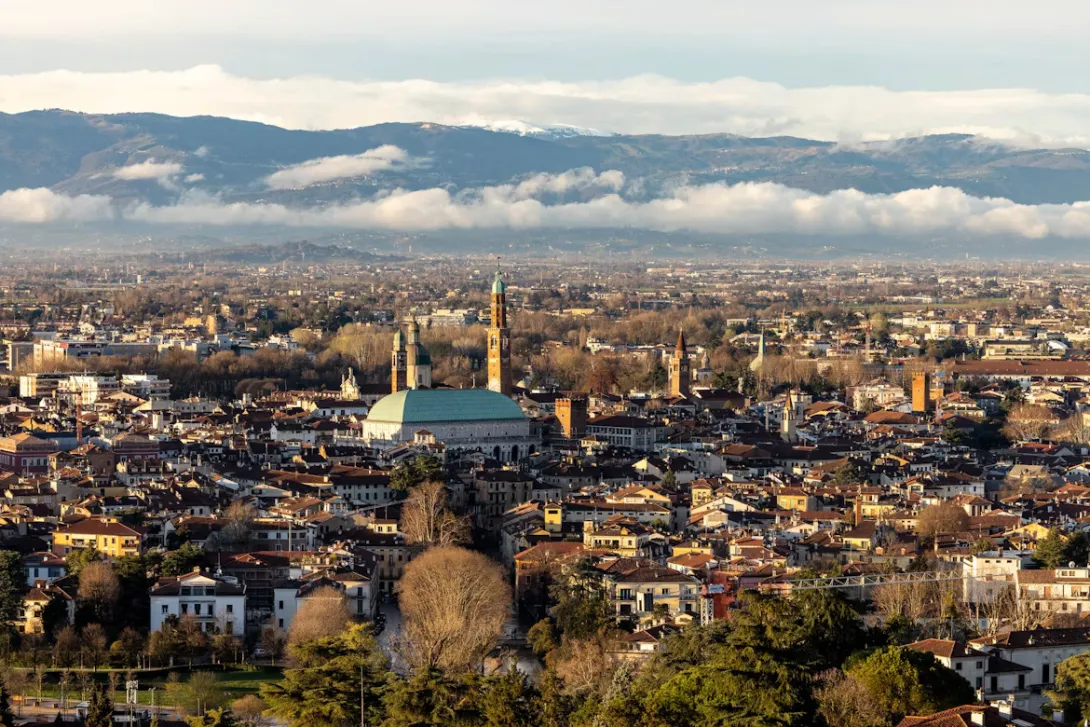 Panoramic view of Vicenza, Italy, featuring elegant Palladian architecture, historic piazzas, church domes, and the surrounding Veneto landscape beneath a clear blue sky.