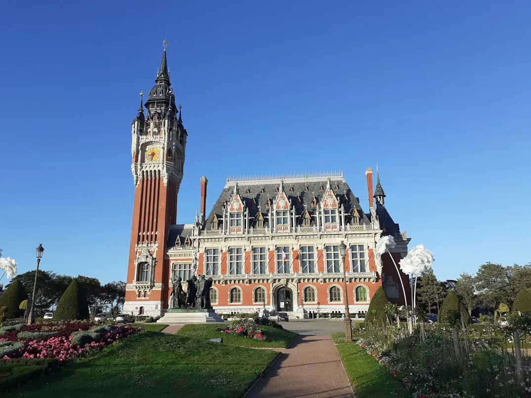 Panoramic view of Calais, France, featuring the historic Town Hall with its Flemish-style belfry, the Channel waterfront, colorful marina, and the famous Burghers of Calais sculpture beneath a bright coastal sky.