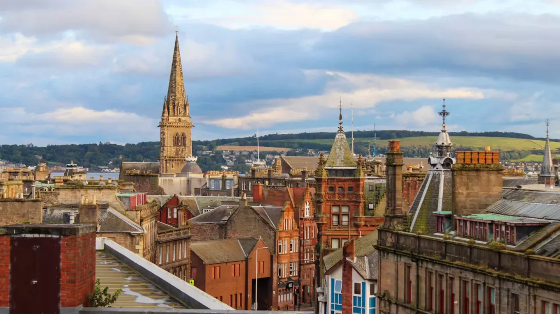 Panoramic view of Dundee, Scotland, featuring the River Tay waterfront, the V&A Dundee museum, historic city buildings, the Tay Bridge, green hills, and the coastal landscape under a bright Scottish sky.