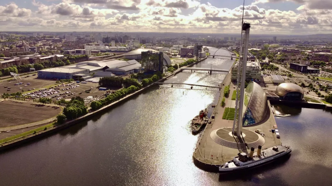 Panoramic view of Glasgow, Scotland, featuring the River Clyde, the city's modern and historic skyline, Victorian architecture, iconic bridges, lush riverside parks, and surrounding green hills beneath a bright Scottish sky.