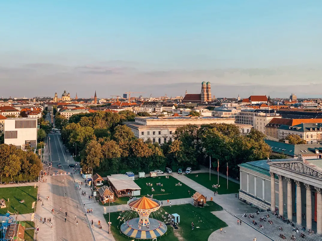 Panoramic view of Munich, Germany, featuring the historic Marienplatz with the Neues Rathaus, Gothic architecture, city rooftops, church towers, green parks, and the urban skyline of Bavaria’s capital under a clear blue sky.