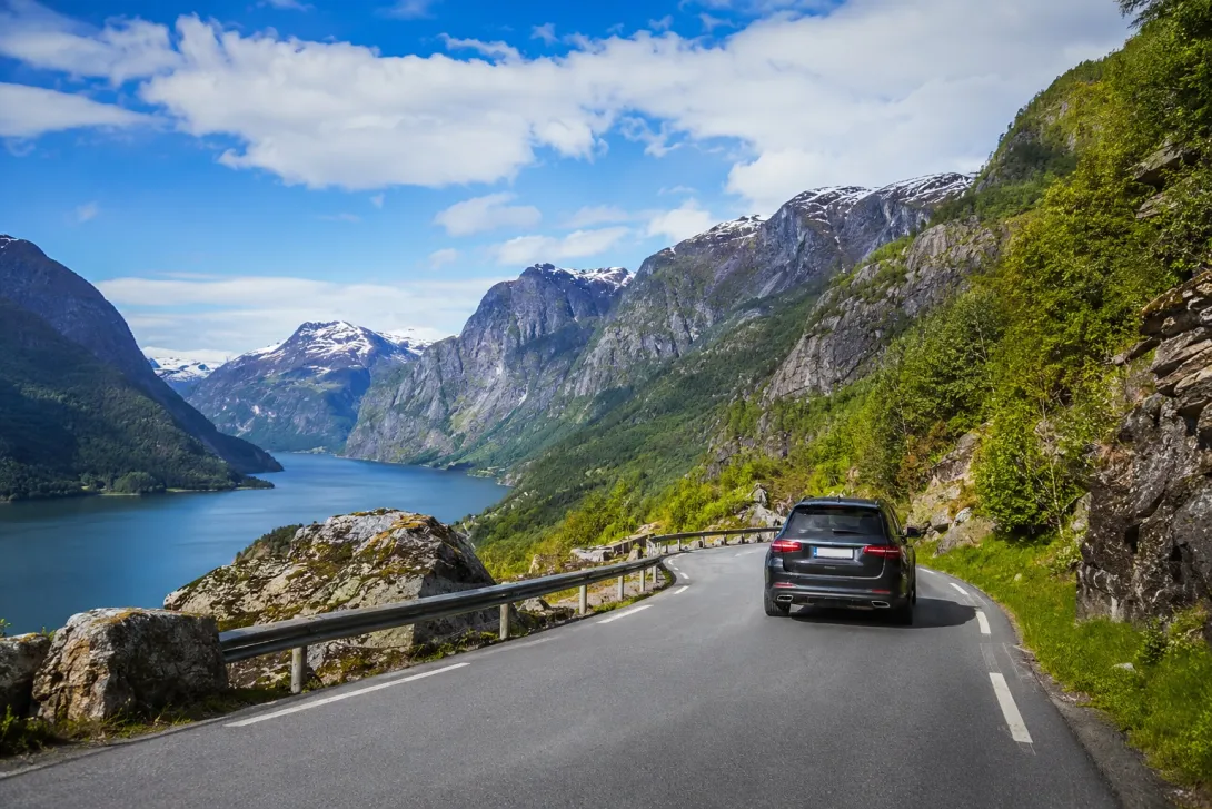 Car driving on a scenic mountain road between Oslo and Flåm, overlooking a Norwegian fjord with snow-capped mountains and dramatic landscapes.