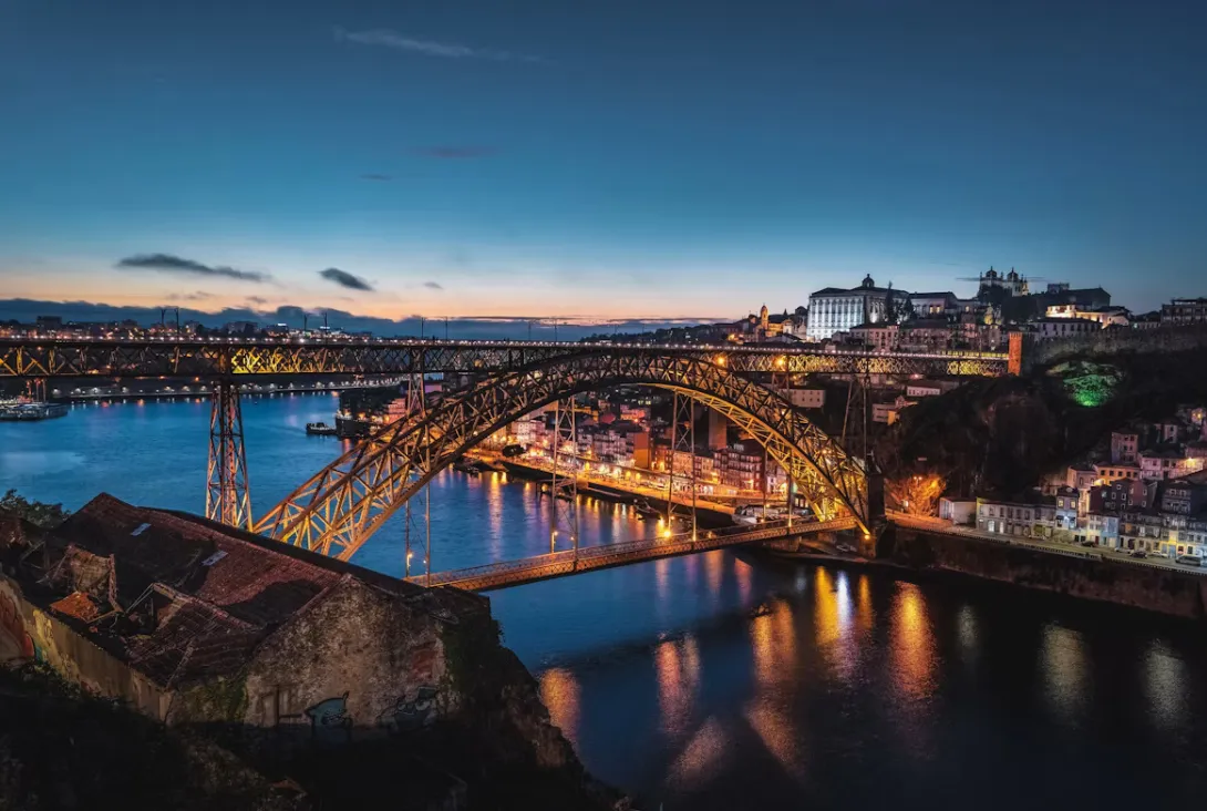 Panoramic view of Porto, Portugal, featuring the colorful Ribeira district along the Douro River, the Dom Luís I Bridge, historic terracotta rooftops, traditional riverside buildings, and the city's iconic skyline under a clear blue sky.