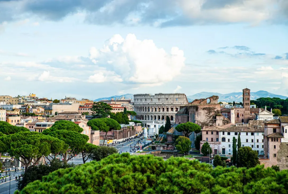 Panoramic view of Rome, Italy, featuring the historic city skyline with the Colosseum, ancient Roman architecture, terracotta rooftops, iconic domes, green parks, and the timeless atmosphere of the Eternal City under a bright Mediterranean sky.