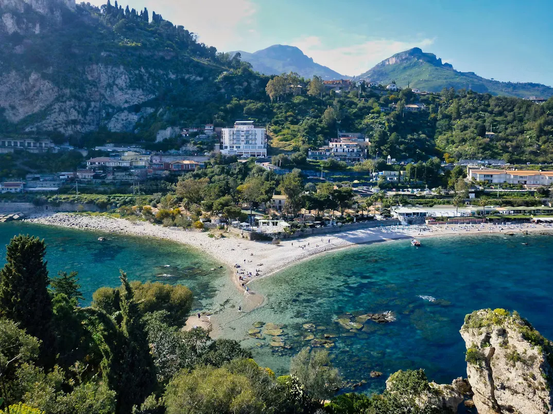 Panoramic view of Taormina, Sicily, featuring the hilltop historic center, the Ancient Greek Theatre overlooking the Ionian Sea, colorful Mediterranean rooftops, lush vegetation, and Mount Etna rising in the background under a clear blue sky.
