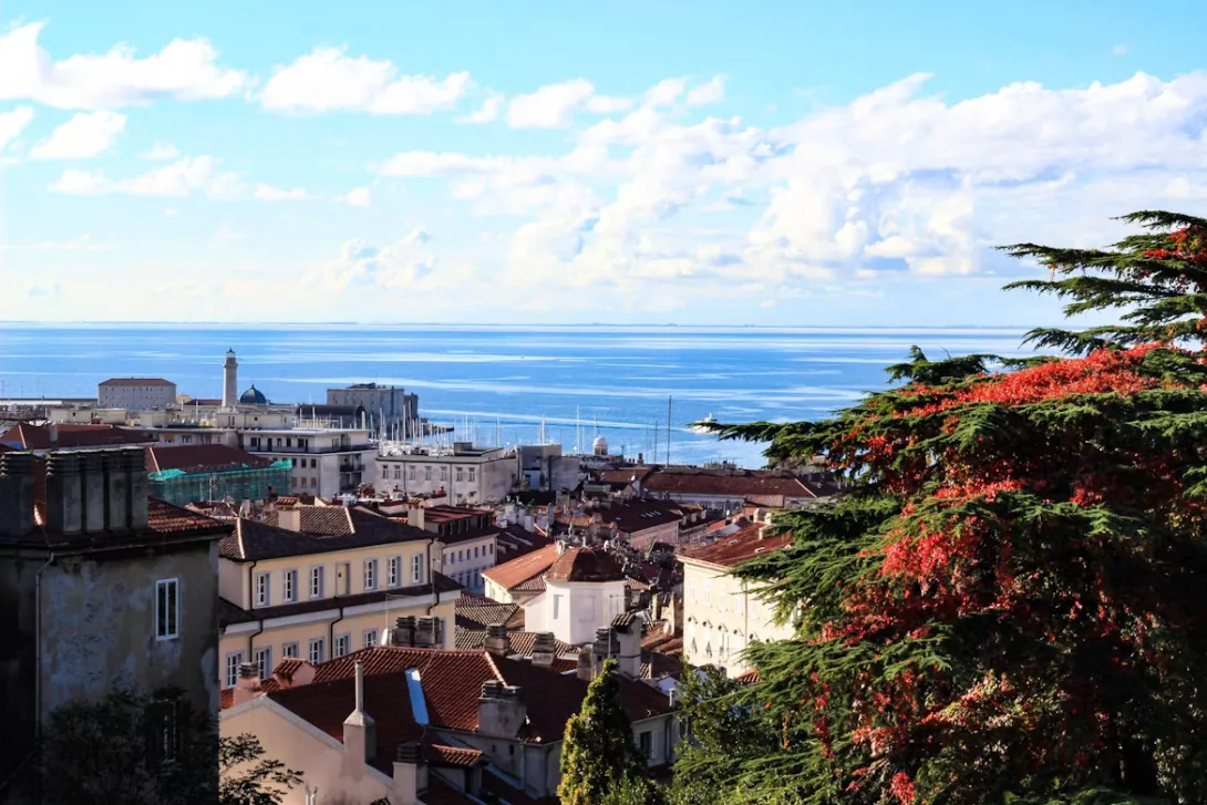 Panoramic view of Trieste, Italy, featuring Piazza Unità d'Italia, the Adriatic waterfront, elegant historic architecture, and Miramare Castle overlooking the Gulf of Trieste on a clear Mediterranean day.