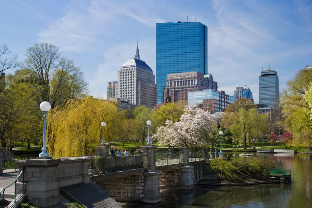 Boston Public Garden with a stone bridge, blooming trees, and the downtown skyline on a sunny spring day, highlighting a popular stop on a Washington, DC to Boston road trip.